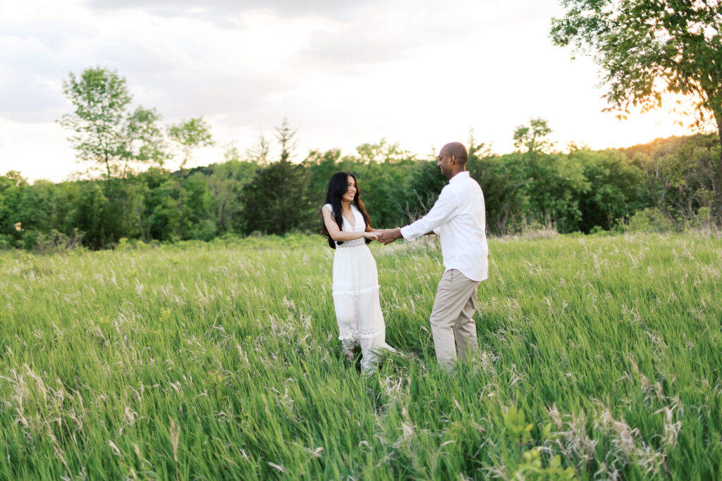 They laugh and dance in the tall grass at sunset, spinning in a circle while holding hands, filled with pure joy and love.