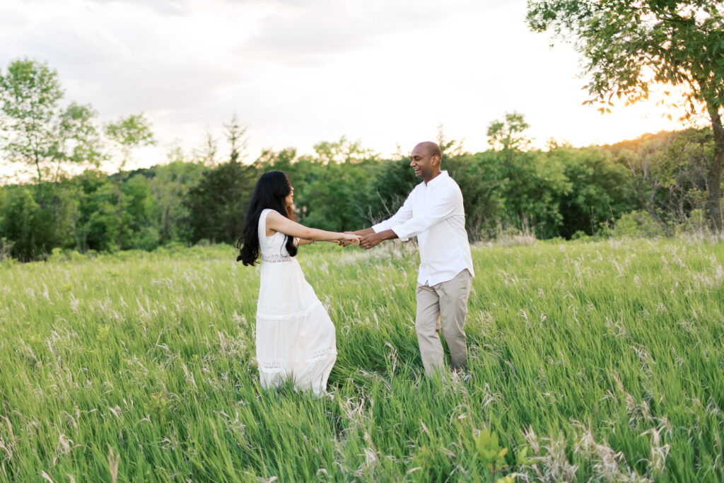 They laugh and dance in the tall grass at sunset, spinning in a circle while holding hands, filled with pure joy and love.