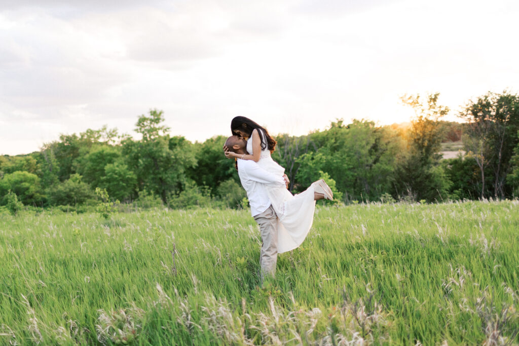 Groom lifts bride during engagement session for a kiss in the warm sunset light, set against a lush green field with trees at Elm Creek Park Reserve in Maple Grove.