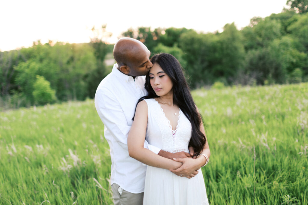 A gentleman kisses his girl on the head while hugging her from behind, capturing a tender moment of love and intimacy. Maple Grove Engagement session.