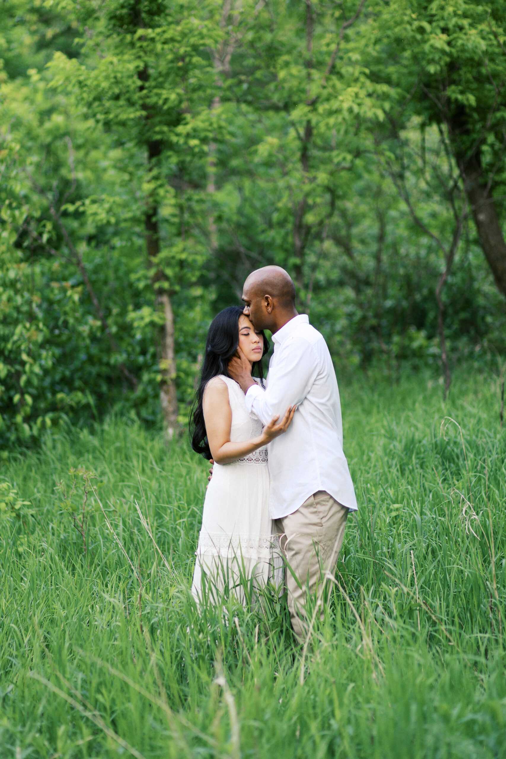 Dominic kissing his bride to be gently in the head. They are surrounded by beautiful green nature in white wardrobes. This is Elm Creek Park in Maple Grove.
