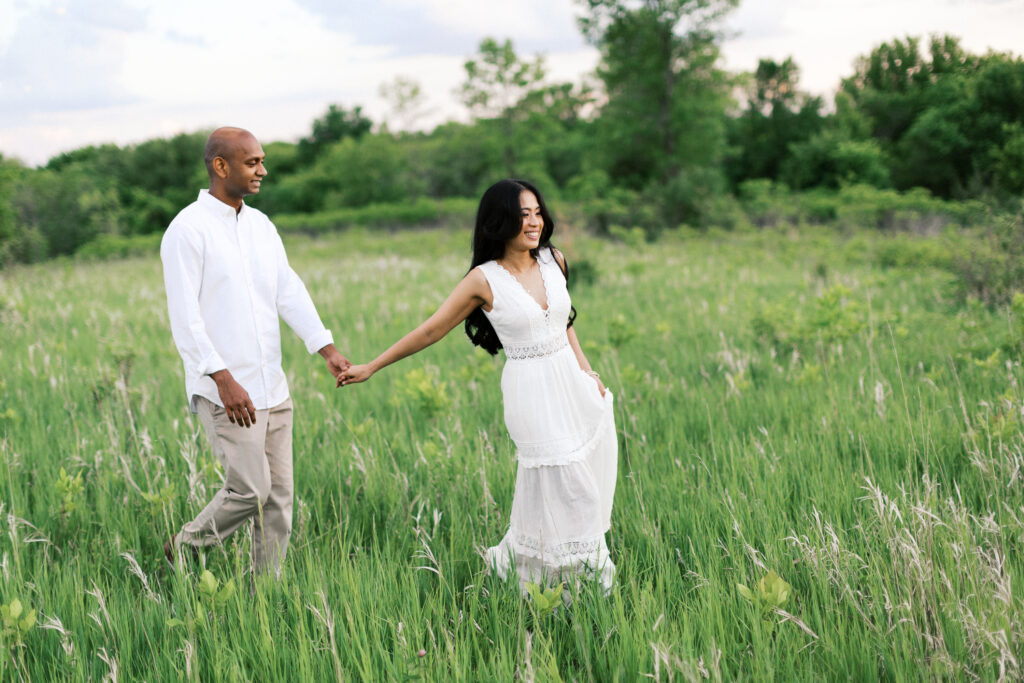 Maple Grove Engagement session. Man and woman walking through a field of tall grass in Maple Grove, holding hands and smiling.
