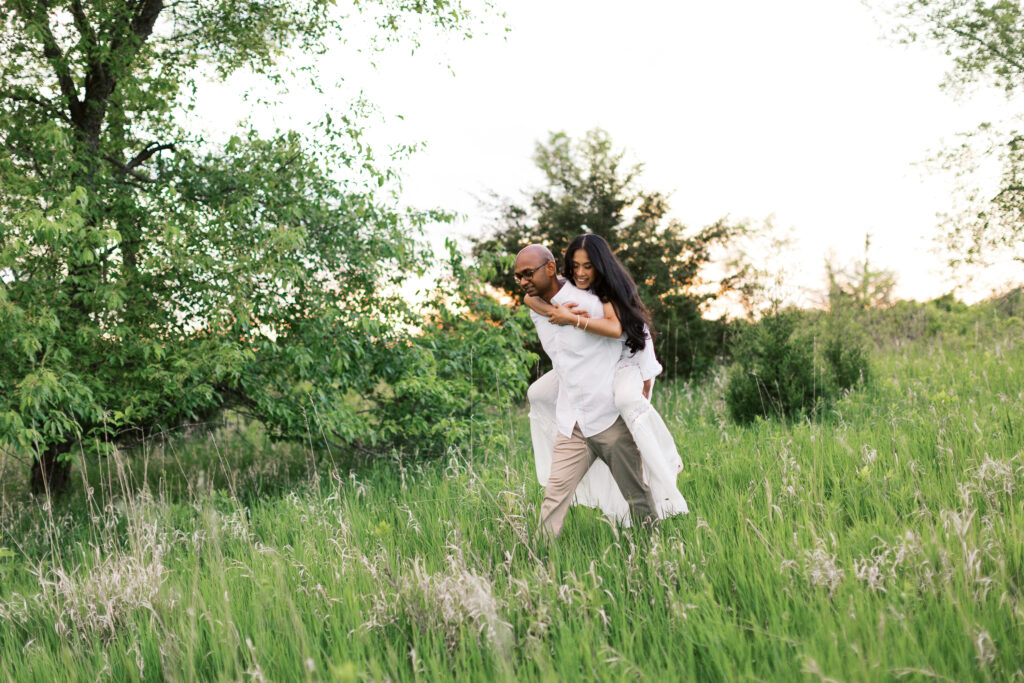 Man gives his partner a playful piggy ride through lush grass as they both smile at Elm Creek Park Reserve in Maple Grove.
