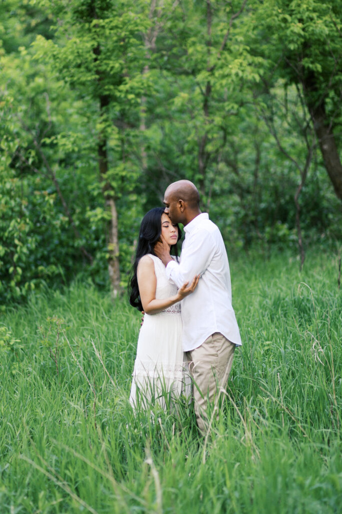 He kisses her head and gently holds her face, standing together in the tall grass, capturing a tender moment of love and intimacy.