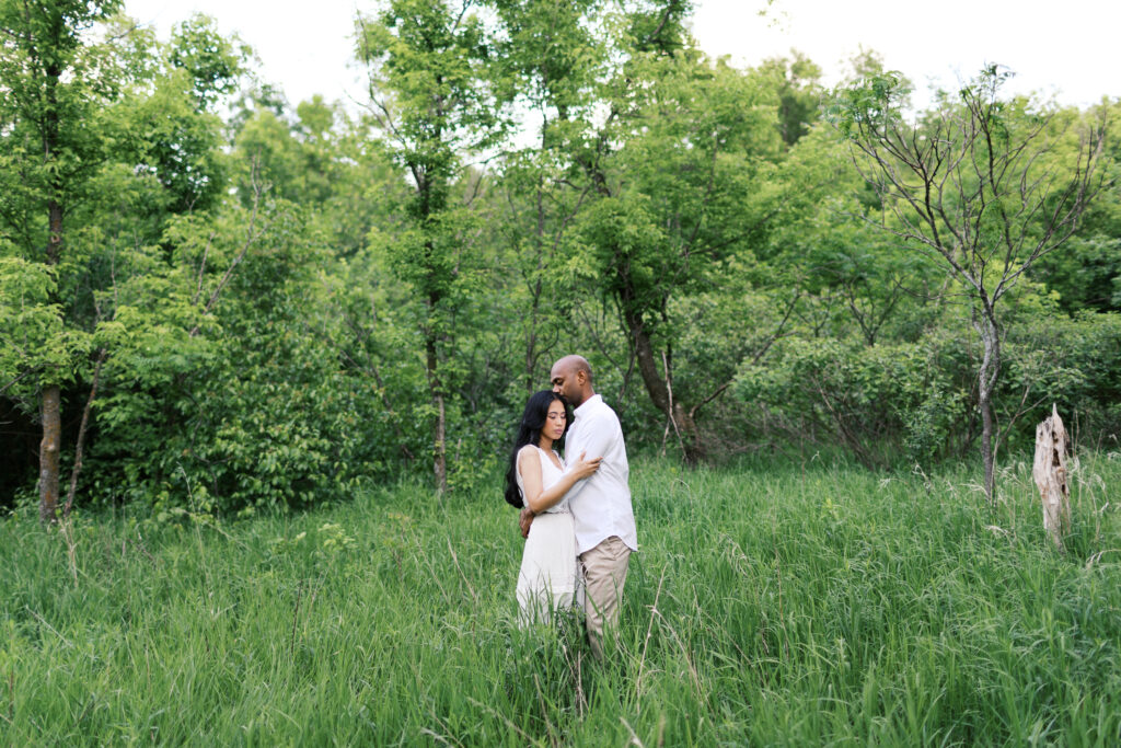 A fine art image of a couple standing in the grass, embracing as he kisses her head with their eyes closed, captured in a candid and intimate moment in nature.