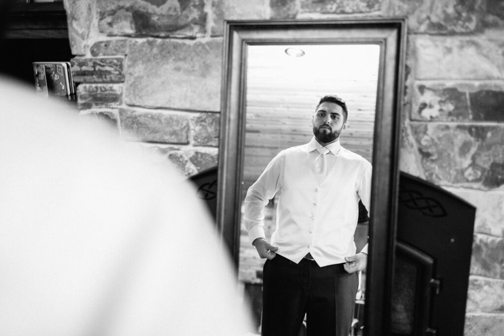 Groom getting ready for his Elopement and looking in the mirror adjusting his white vest.