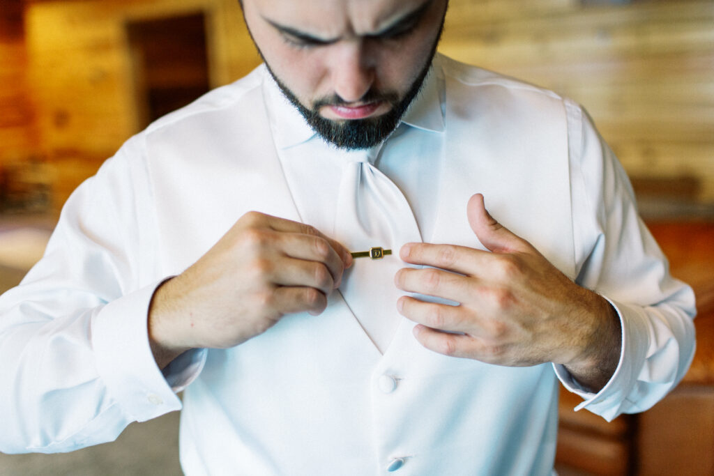 Groom putting on a gold tie pin to hold his tie in place for his wedding in Webster Wisconsin. Captured ny Minnesota wedding photographer, Toly Dzyuba.