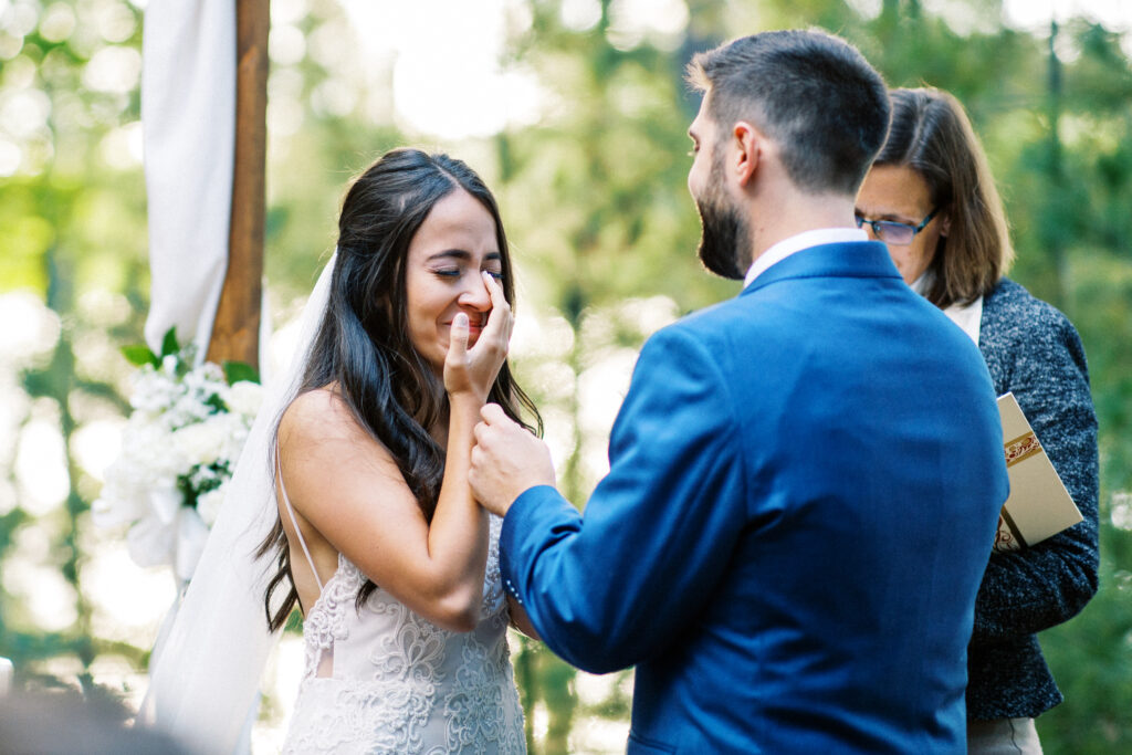 Bride is crying with tears of joy at her wedding ceremony while groom gently helps to wipe her tears away. A Story of Love: Priscilla & Jack’s Very intimate moment at the cabin.