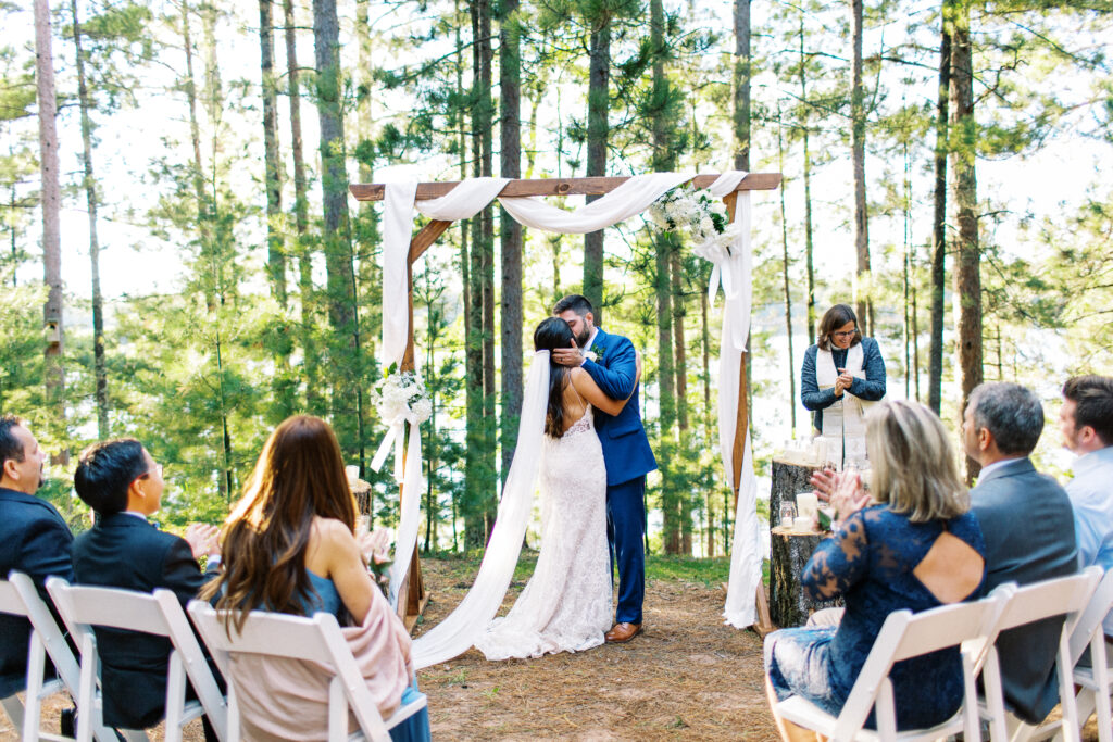 Bride and groom kissing at the altar surrounded by nature on their wedding day, with the sound of cheers from family.