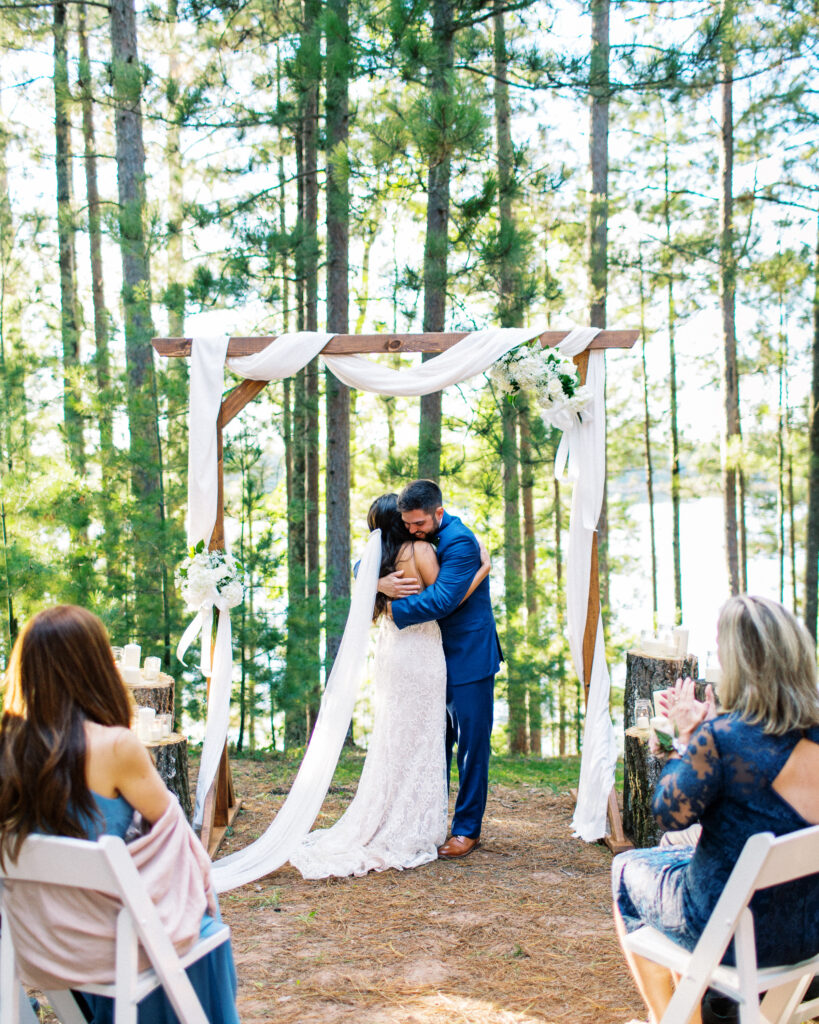 Bride and groom hug and kiss at the wedding ceremony as sunlight sets with guests clapping with cheers. This scene occurred during a heartwarming cabin elopement in Webster, WI captured by a Minnesota Wedding Photographer.