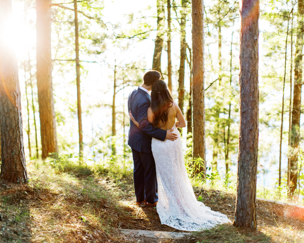 A heartfelt moment as bride and groom hugs in the woods, looking at the lake and sun rays beaming through the woods. This scene occurred during a heartwarming cabin elopement in Webster, WI captured by a Minnesota Wedding Photographer.
