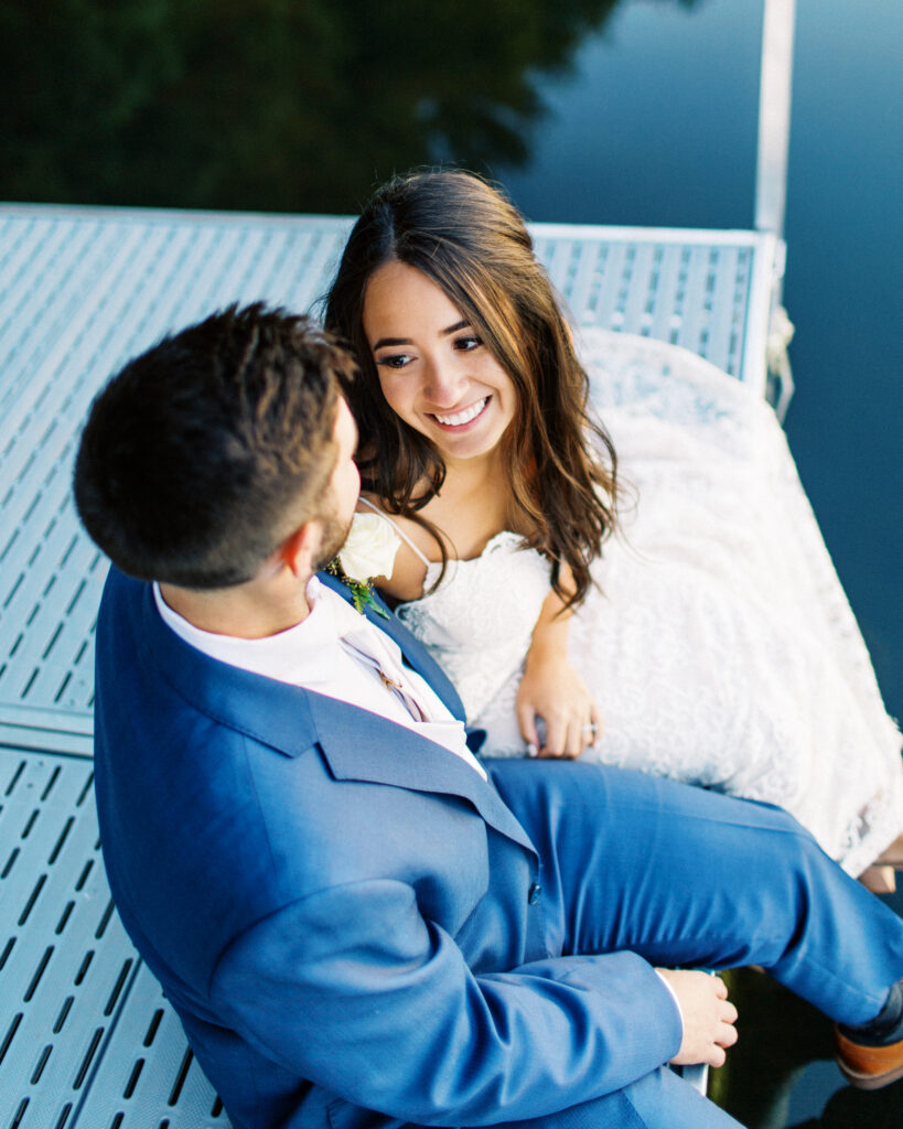 Bride and groom sitting on the dock surrounded by peaceful water looking at each other and smiling. A true heartfelt Cabin Elopement in Webster, WI | Minnesota Wedding Photographer.