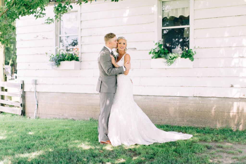 Groom gently kisses his bride’s forehead as she looks away, standing by the old white cottage house at Legacy Hill Farm. Captured by Minnesota wedding photographer.