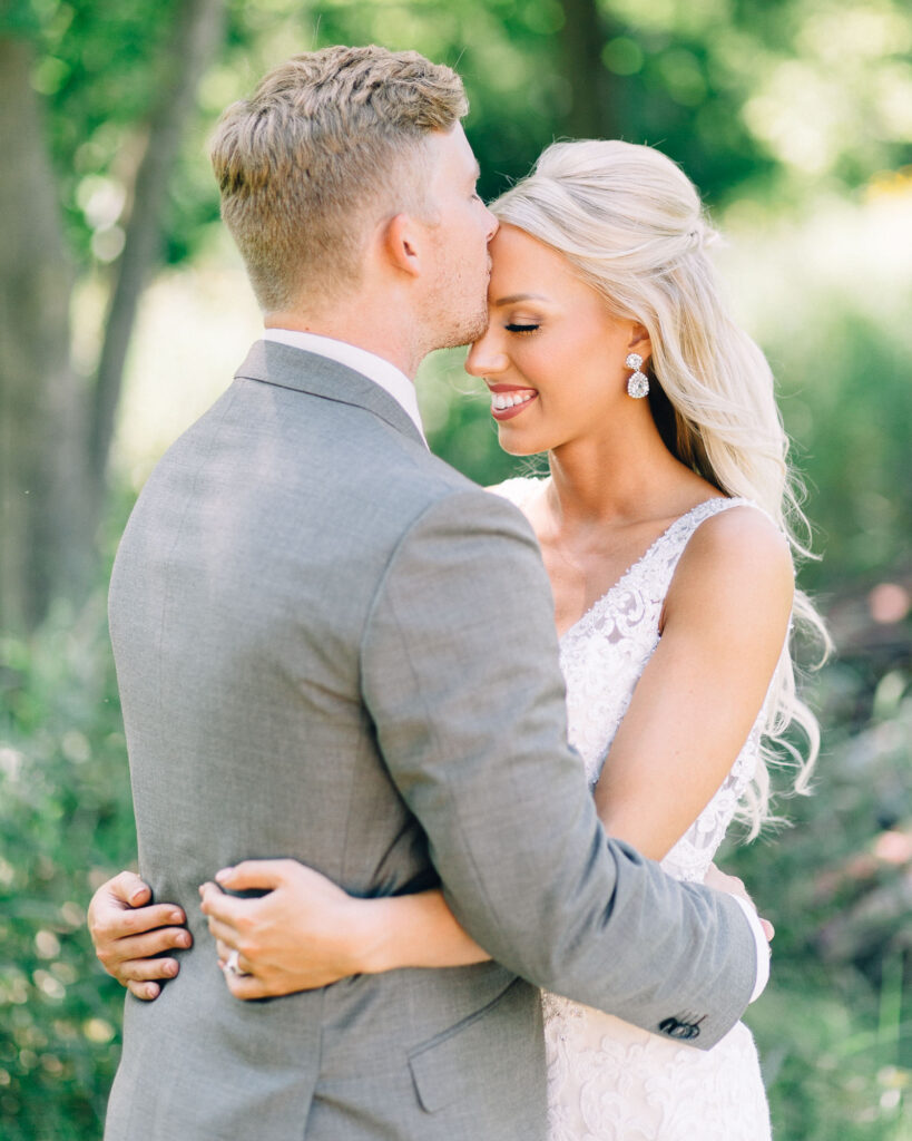 Groom kisses his bride in the forehead while she closes her eyes with a smile.