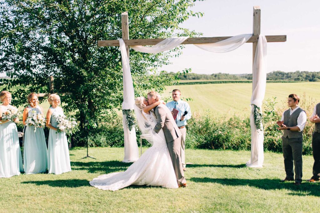 Groom hugs and kisses his bride during the wedding ceremony, overlooking the farm fields at Legacy Hill Farm, captured by Toly Dzyuba Photography.