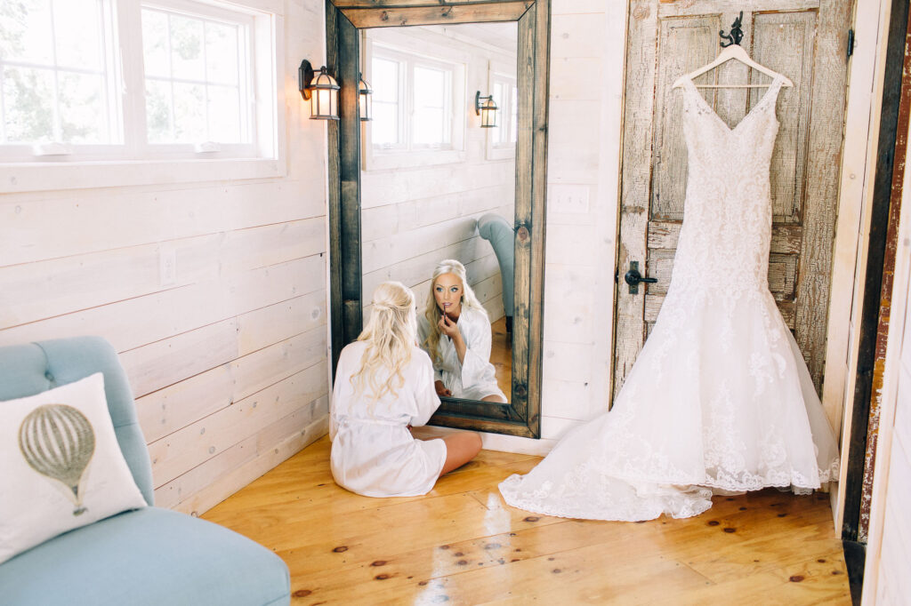Bride in her rope sitting at the mirror on the floor and putting on her lipstick, with the dress hanging next to her on the rustic door. This is at Legacy Hill Farm.