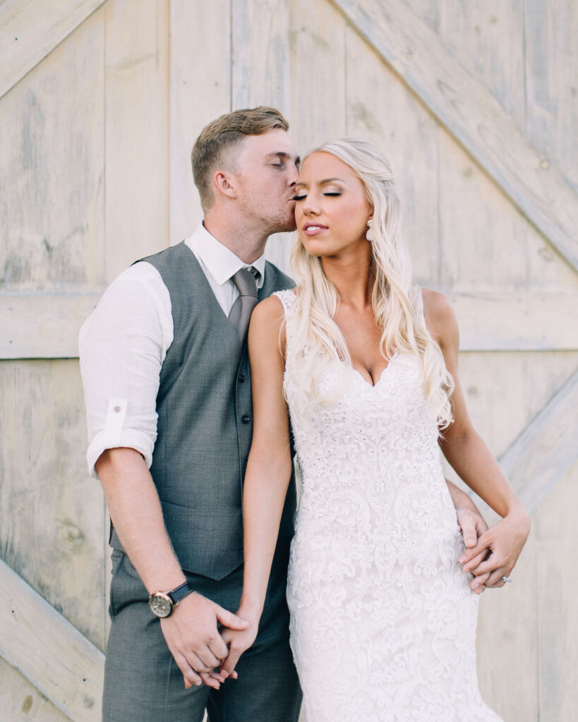 Bride and groom stand together at Legacy Hill Farm, with the groom kissing her head as she closes her eyes, holding hands in a romantic moment, captured by Minnesota wedding photographer Toly Dzyuba.