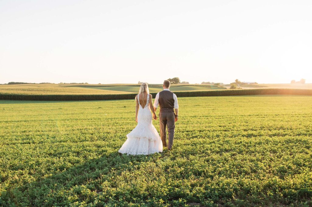 Bride and groom holding hands, walking across the farm field at Legacy Hill Farm. Warm, romantic light beams over them as they stroll, captured by Toly Dzyuba Photography.
