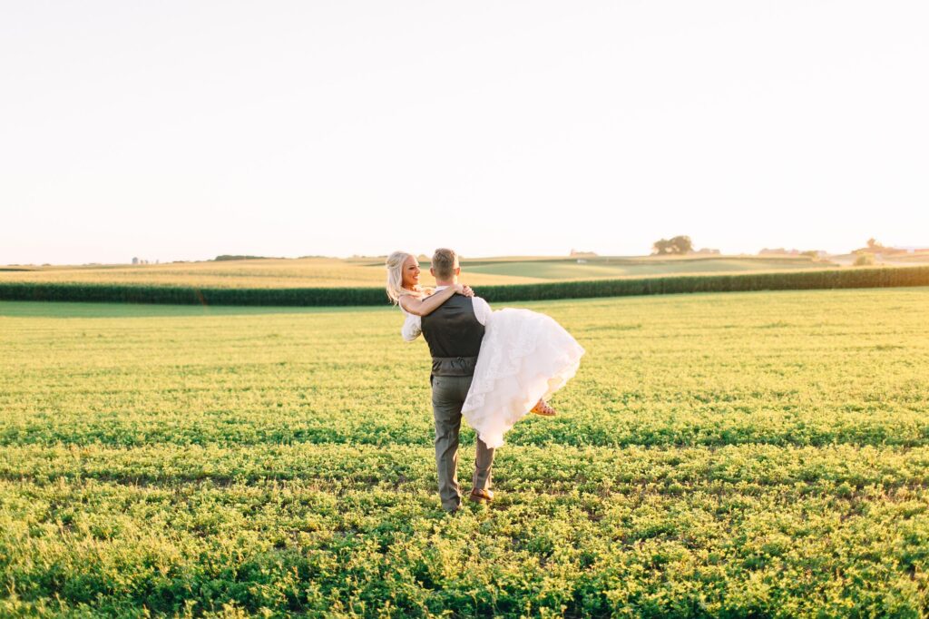Groom picks up his bride, their heads touching, as he carries her across the field at Legacy Hill Farm with a sunset backdrop and romantic vibes, captured by Toly Dzyuba Photography.