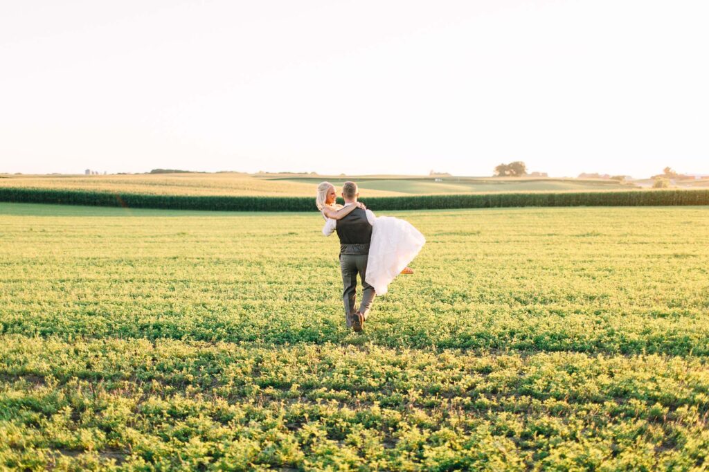 Groom carries his bride across the farm field at Legacy Hill Farm, holding her in his arms with a sunset backdrop casting buttery tones, captured by Minneapolis wedding photographer Toly Dzyuba.