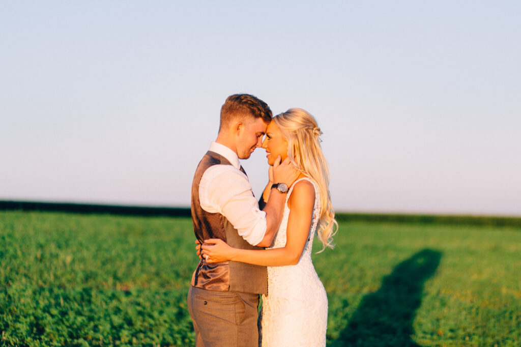 Bride and groom touch heads together, with his hands gently on her face, as they share an intimate moment with the sunset glowing in the background.