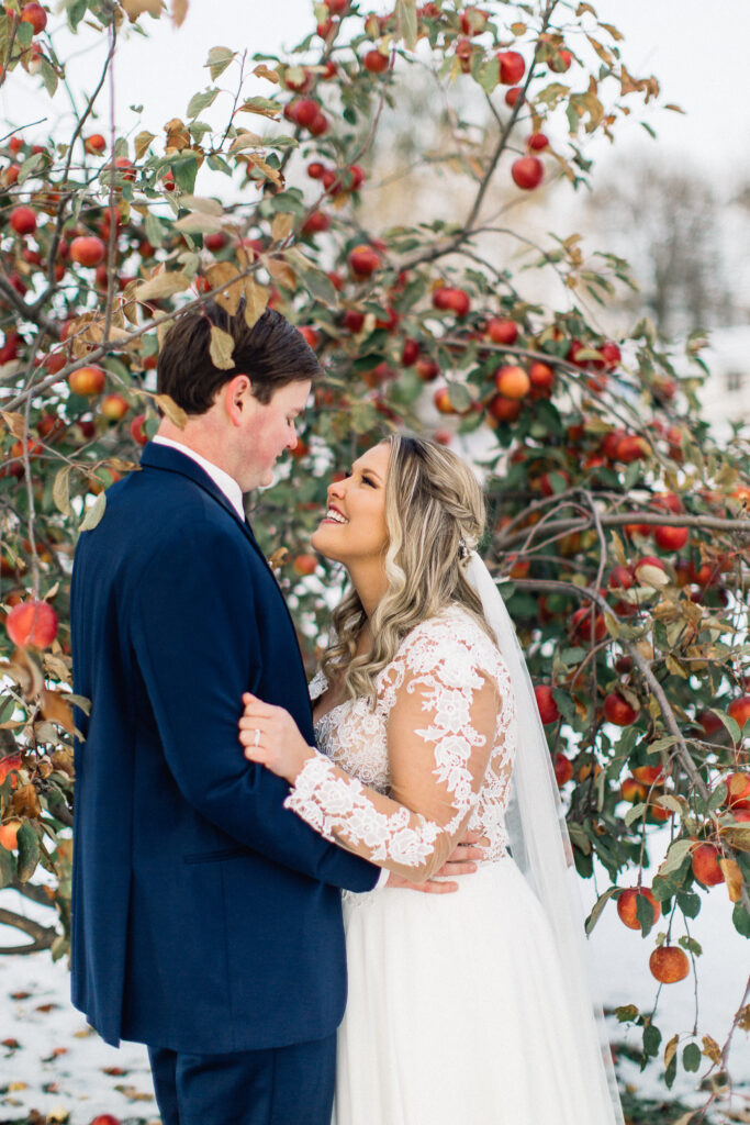 Bride and groom gaze passionately at each other amidst red apples in the background and snow on the ground during their winter wedding at The Cottage Farmhouse.