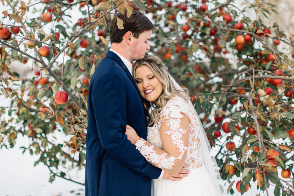 Bride leaning on groom's chest on a wintry cold day with a smile at The Cottage Farmhouse, with an apple tree and red apples in the background.