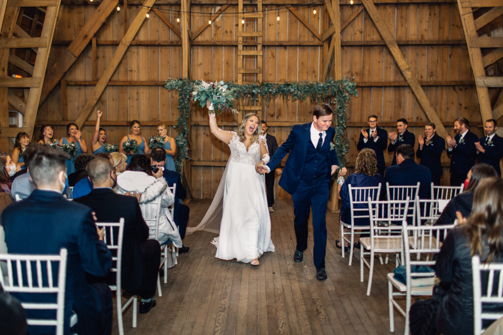 Bride and groom celebrate their marriage as they exit the ceremony, with the bride raising her bouquet and guests cheering, captured by a Minneapolis wedding photographer at The Cottage Farmhouse.