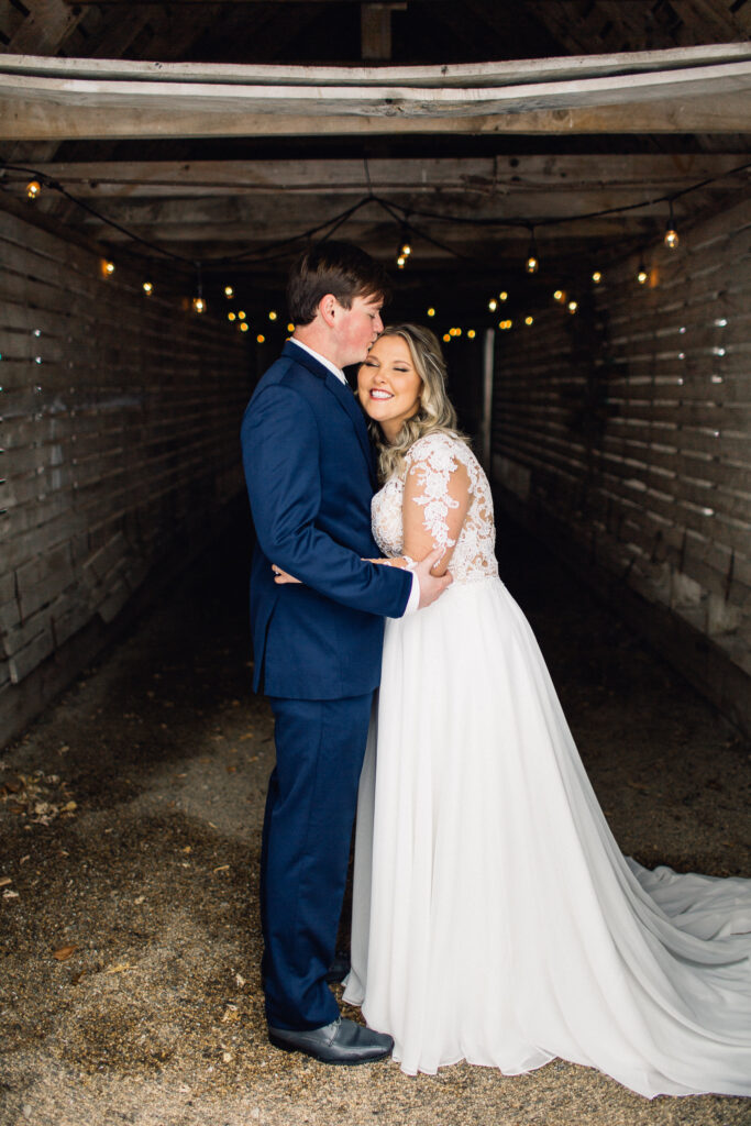 Groom kisses his bride in the head inside a small rustic barn at the Cottage Farmhouse.