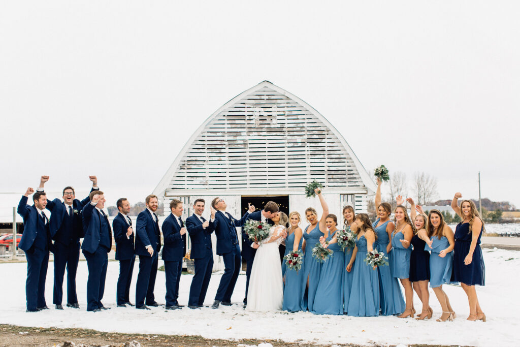Wedding party cheers as bride and groom kiss at The Cottage Farmhouse on a winter day, captured by Minnesota wedding photographer.