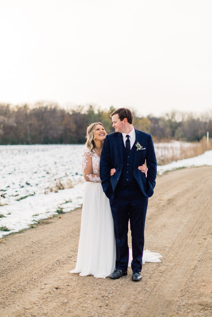 Bride stands behind groom with her arms around his waist as they look at each other during sunset at The Cottage Farmhouse.