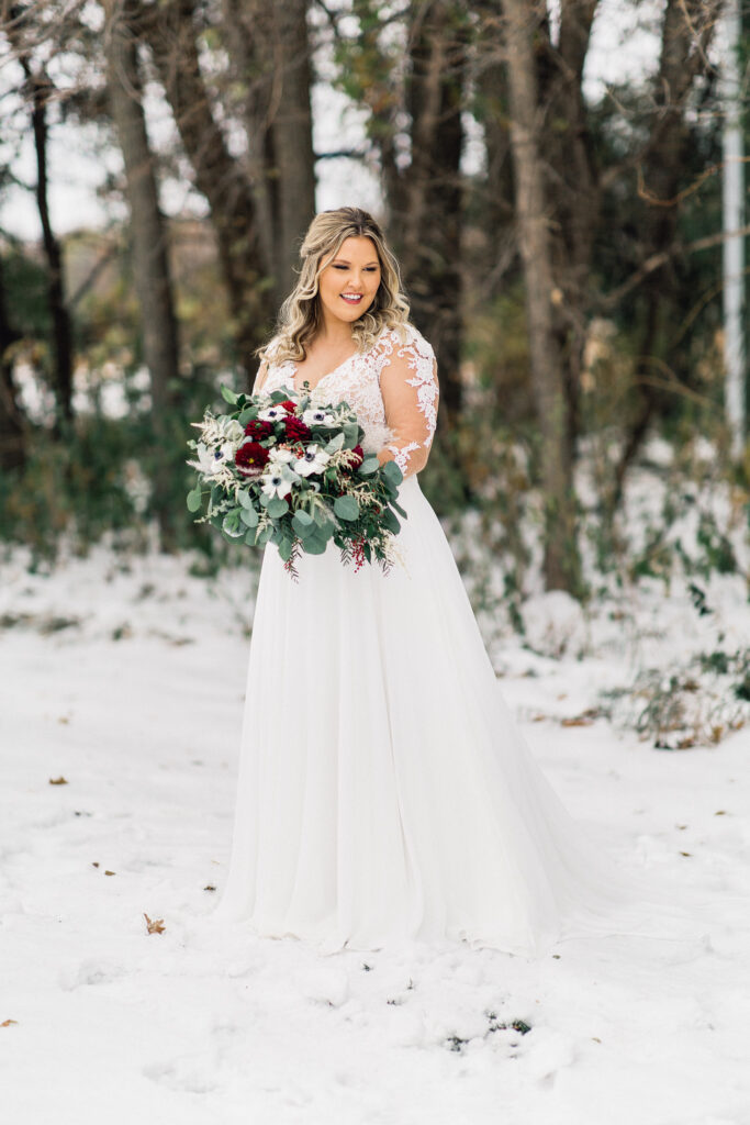 Bride standing tall looks to the side and down with her large beautiful bouquet in her hands and snow on the ground at The Cottage Farmhouse winter wedding.