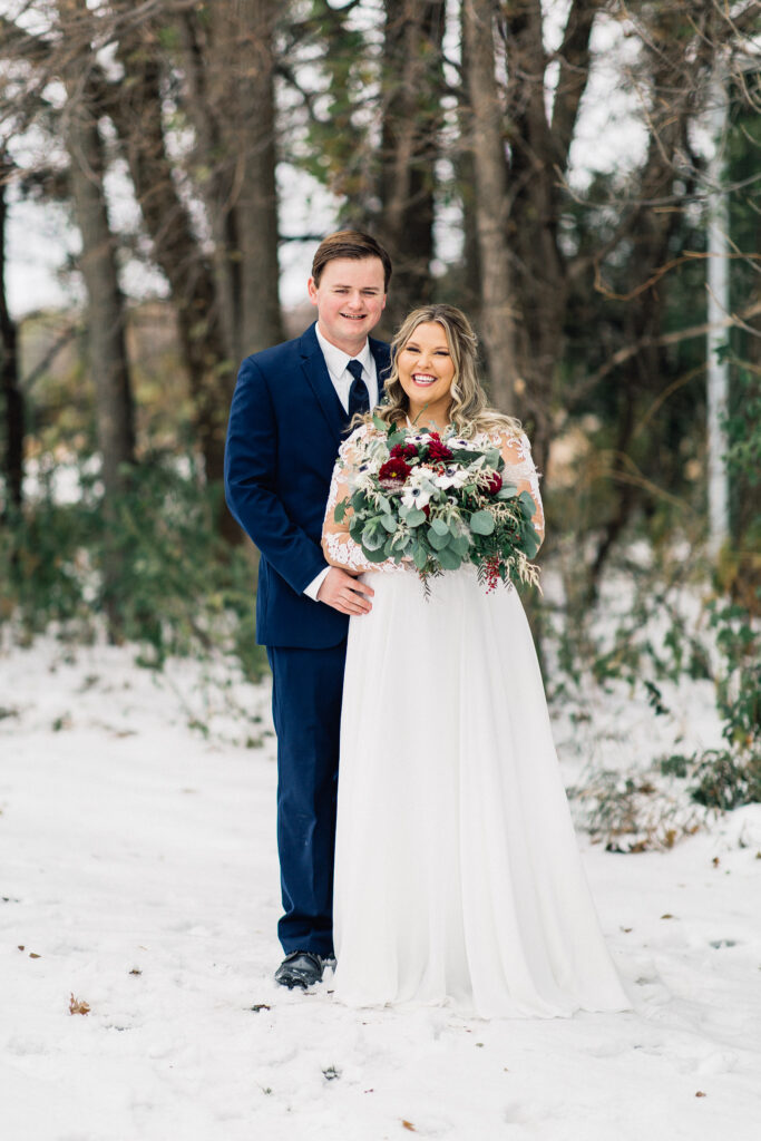 Groom stands behind bride with his arm around her waist, smiling, while she holds her gorgeous bouquet at The Cottage Farmhouse winter wedding.