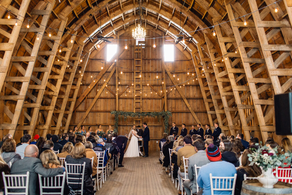 Wedding ceremony in the rustic barn at The Cottage Farmhouse, with guests seated as the bride and groom hold hands, captured by a Minnesota wedding photographer.