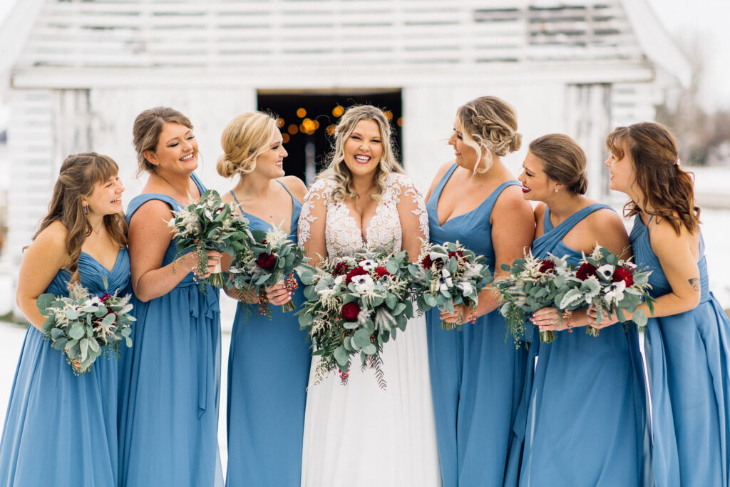 Bridesmaids looking at the smiling bride holding flowers at The Cottage Farmhouse, captured by Minnesota wedding photographer.