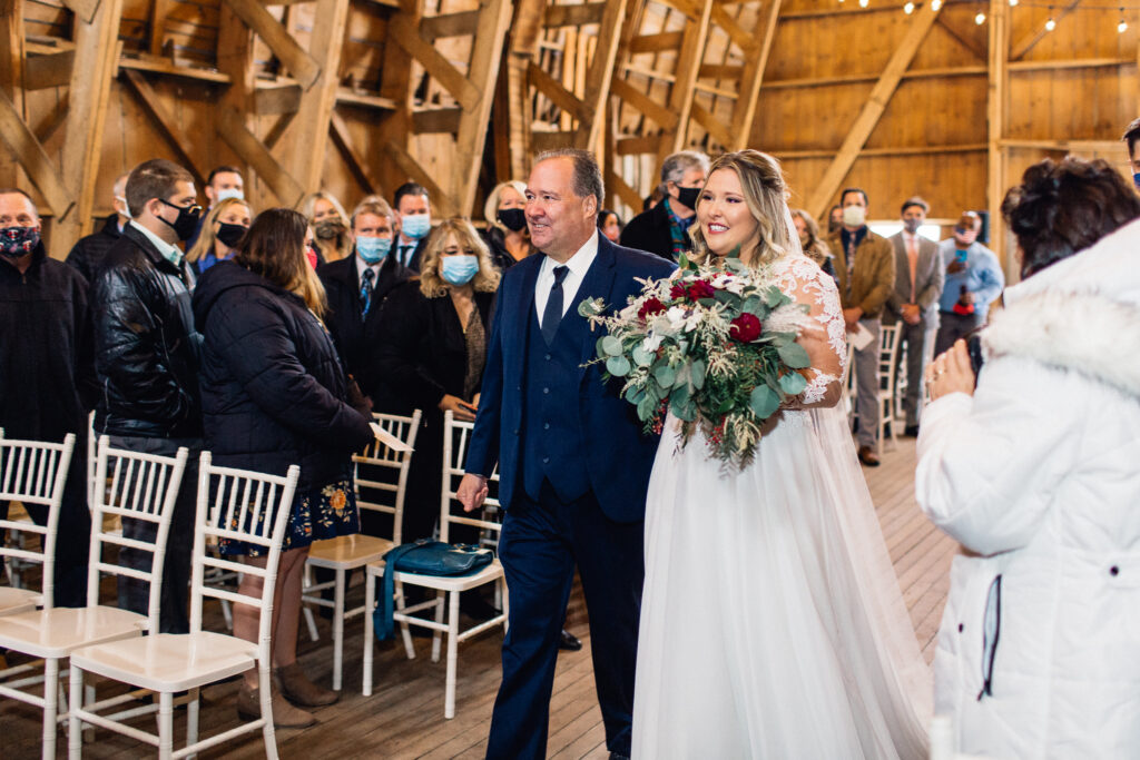 Bride walks down the aisle with her father, both smiling, in the barn at The Cottage Farmhouse.