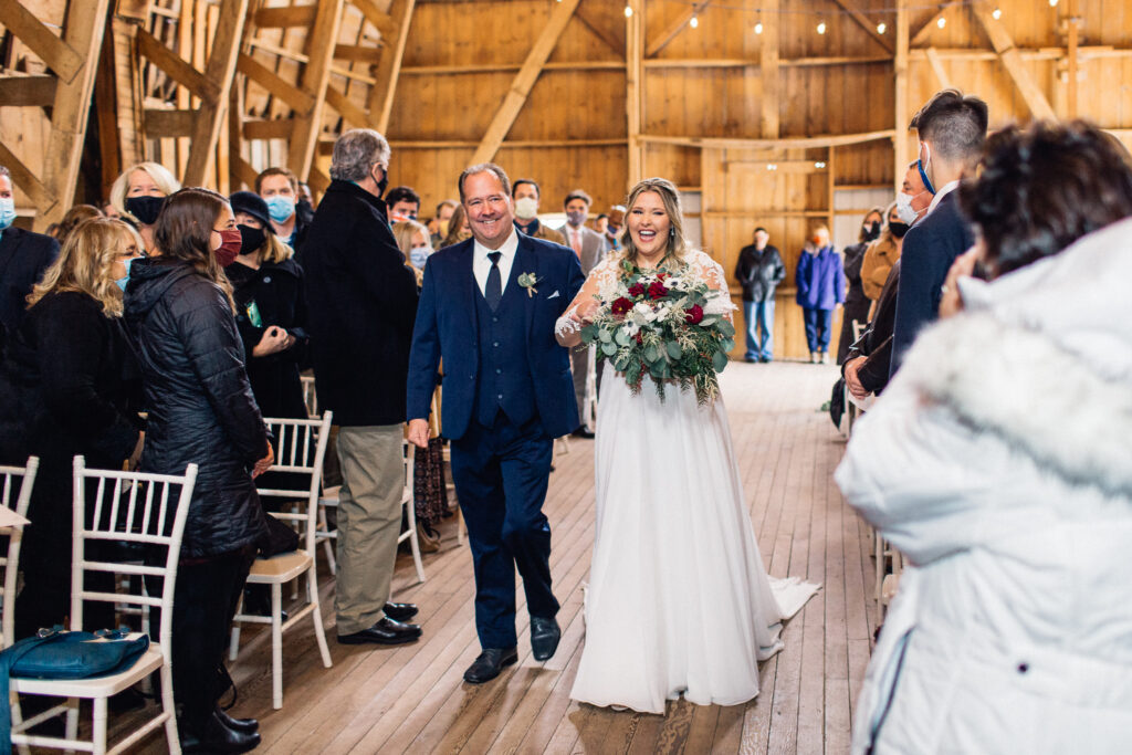 Bride walks down the aisle with her father, both smiling, in the barn at The Cottage Farmhouse.