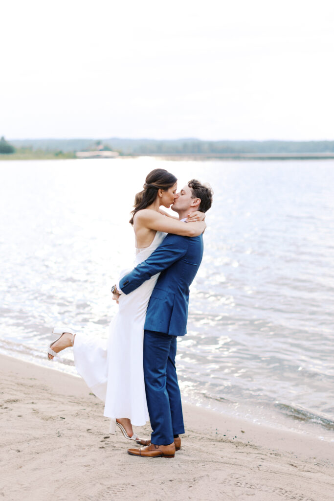 Bride and groom share a kiss on the beach while the groom lifts her during their wedding at Madden’s Resort on Gull Lake, captured by Minnesota wedding photographer.