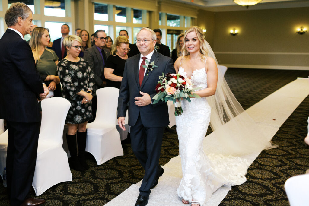 Dani wearing a Ziel Bridal gown walking down the aisle with her father, with joyful guests looking