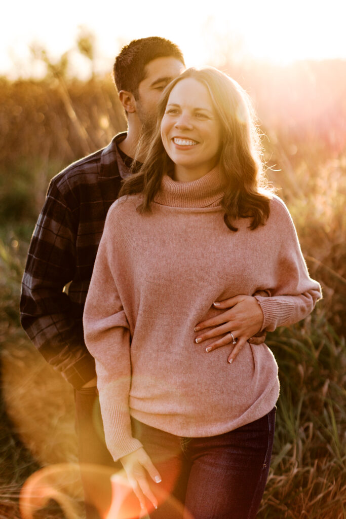 Capturing a moment of pure bliss, this image showcases a tender embrace: he gently stands behind her, his lips brushing her hair as she beams with joy. The warm, golden hues of the setting sun paint the sky, creating a breathtaking backdrop for this intimate scene, all beautifully framed within the enchanting landscape of Elm Creek Park Reserve in Maple Grove. This photograph, skillfully captured by a talented Minnesota wedding photographer, encapsulates the essence of love, serenity, and the magic of a truly unforgettable engagement session.