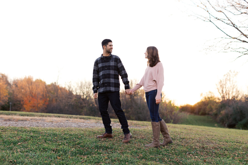 Man holding her hand as they walk across the grass, gazing into each other's eyes at sunset in Elm Creek Park Reserve, Maple Grove, captured by a Minnesota wedding photographer.