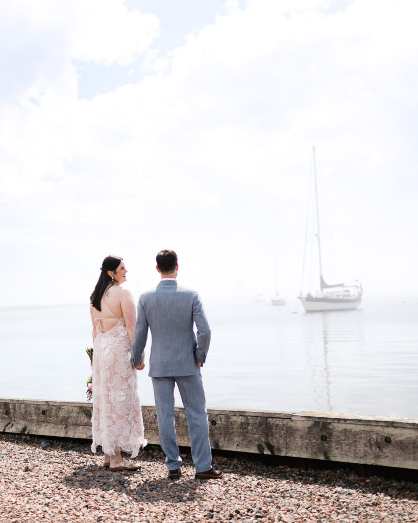 Groom holding his bride on the shoreline of Lake Superior during their North Shore wedding with sailboats on the background. Captured by Grand Marais wedding photographer.