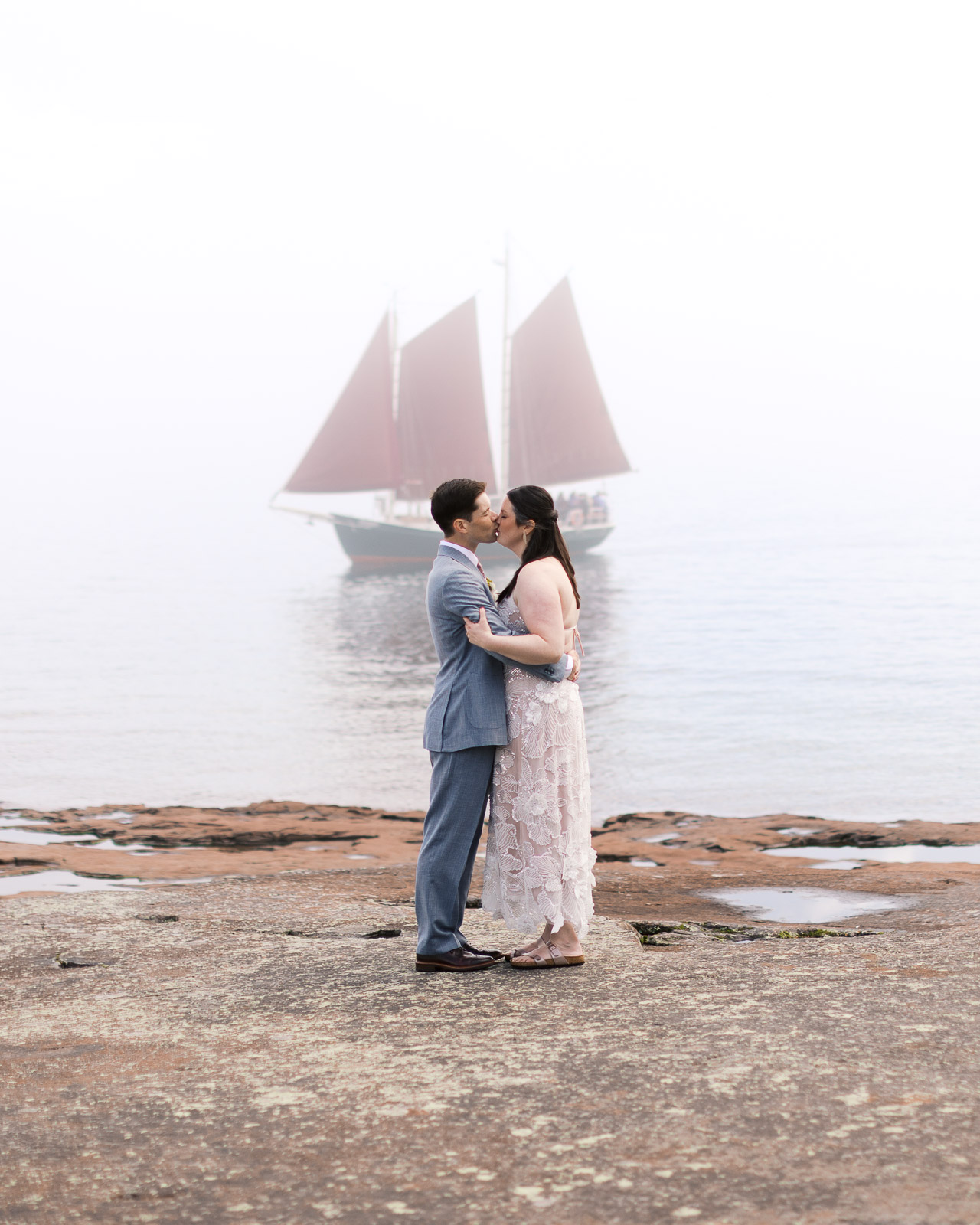 North Shore wedding portrait with breathtaking Lake Superior views and sailboat passing by as the couple kiss at Grand Marais wedding.