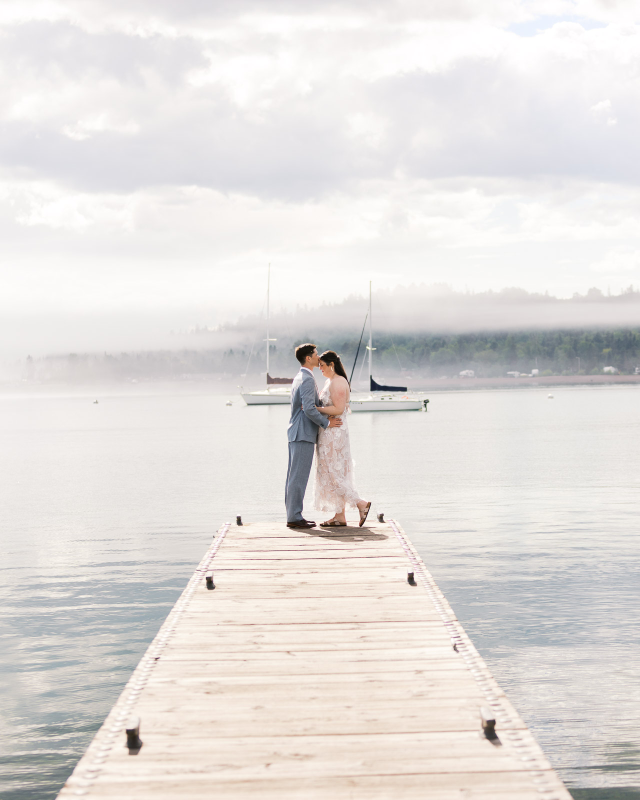Groom kissing his bride on a dock in Grand Marais, Minnesota, with a sailboat in the background, photographed by Toly Dzyuba Photography, Minnesota wedding photographer.